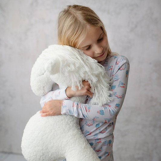 A young girl with blonde hair wearing a light blue and pink unicorn pajama set, hugging a large, fluffy white mermaid-shaped plush pillow with a fringed neck and tail against a light gray textured wall.