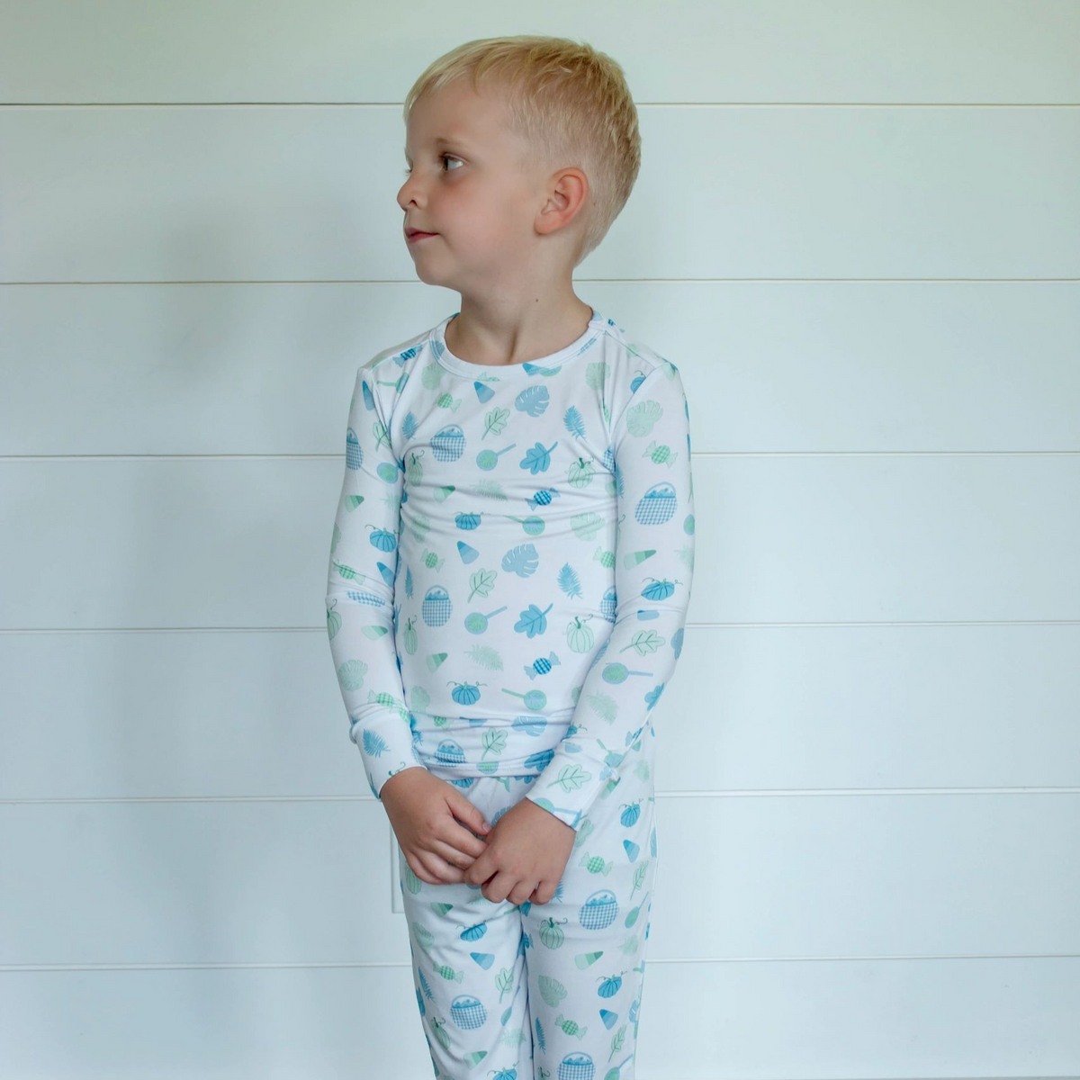 A young child wearing a white long-sleeve pajama set with a light blue cloud pattern, standing against a white paneled wall.