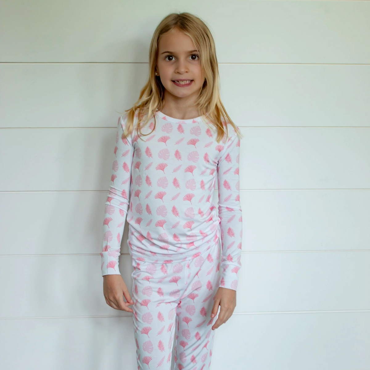 A young girl stands barefoot against a white shiplap wall, wearing a matching long-sleeve pajama set with a pink and white seashell pattern.