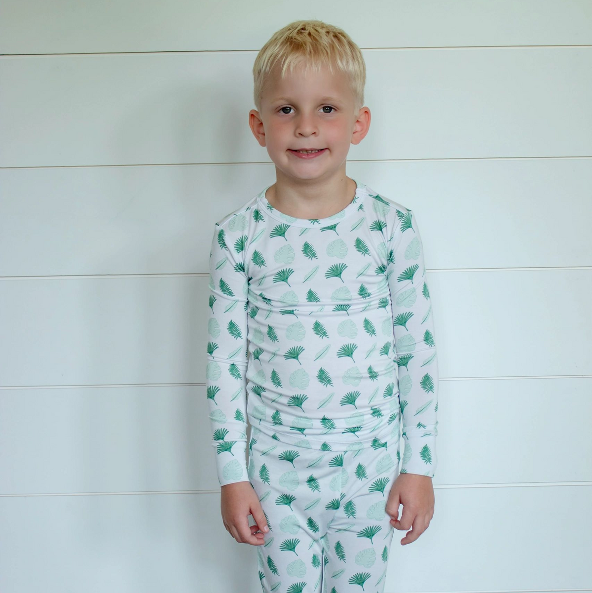 A young boy with blonde hair stands against a white shiplap wall wearing a white long-sleeve pajama set with a green dinosaur pattern.
