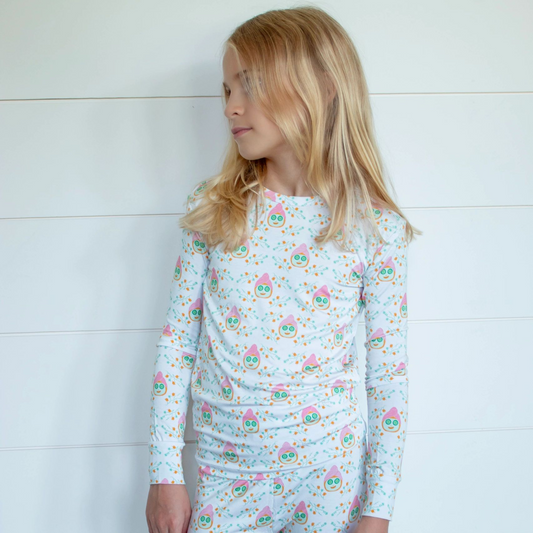 A young girl with blonde hair stands against a white paneled wall wearing a matching long-sleeve top and pants pajama set with a colorful floral pattern.